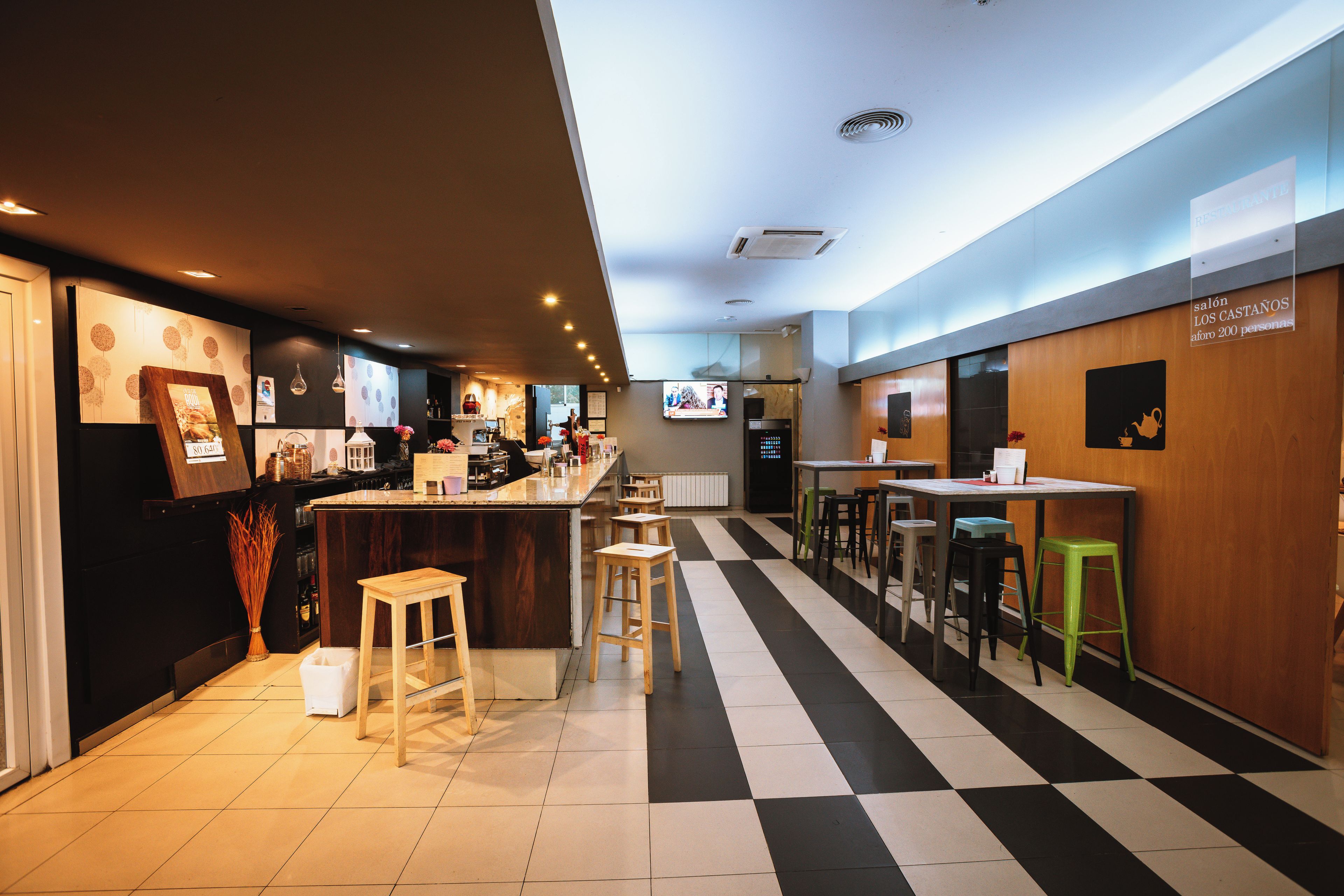 Un restaurante con suelo a cuadros, barra, taburetes y una pantalla de televisión.