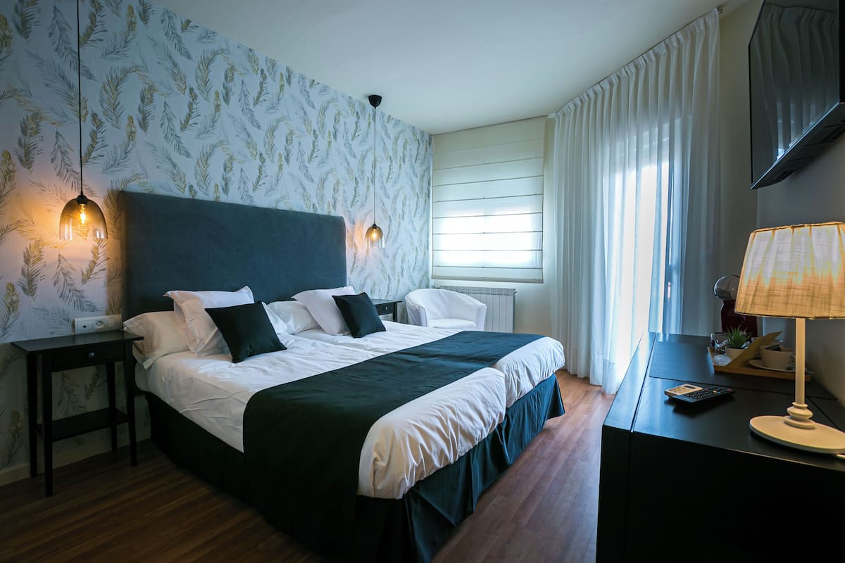 A neatly made bed with a dark headboard, a bedside table with a lamp, and a window with curtains.