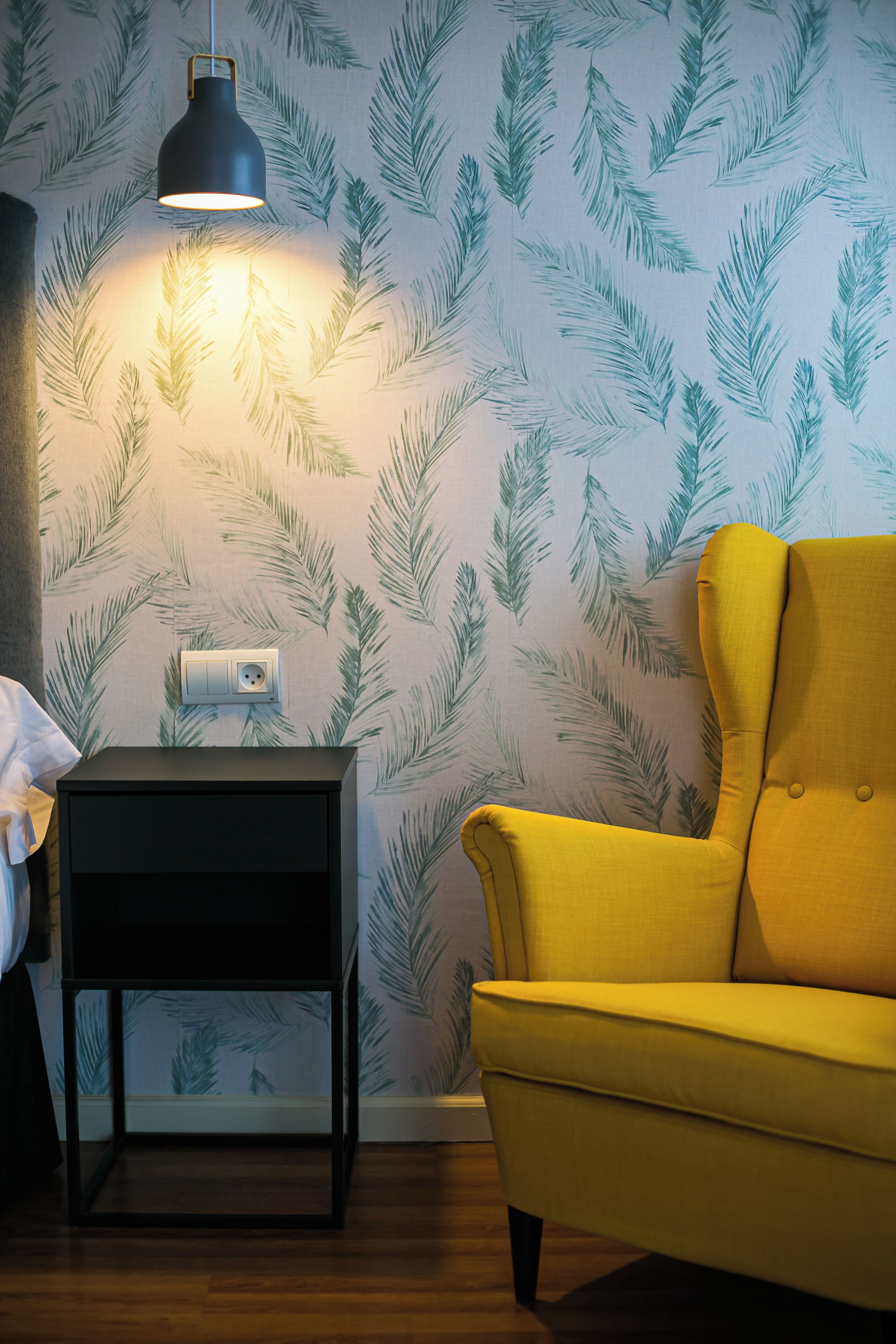 Una habitación con papel tapiz estampado, un sillón amarillo, una mesita de noche negra y una lámpara colgante.