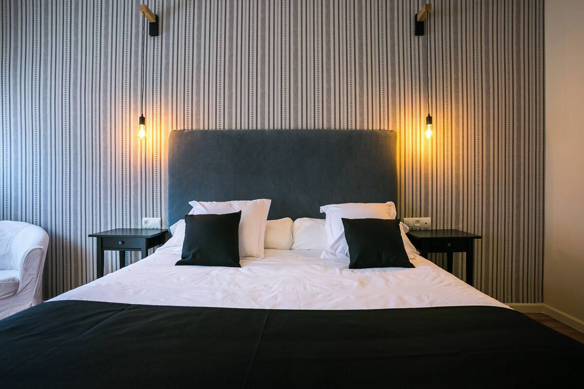 A neatly made bed with white and black bedding, two bedside tables with lamps, and a striped wallpaper background.
