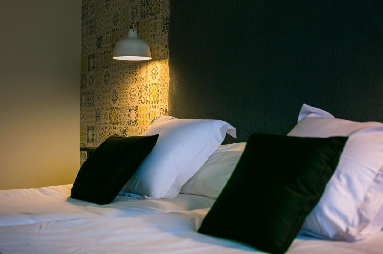 Ein ordentlich bezogenes Bett mit weißen und schwarzen Kissen, einem gemusterten Wandpapier und einer Wandlampe.