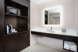 A modern bathroom with a dark wood vanity, a microwave, a coffee maker, and a large mirror with built-in lighting.
