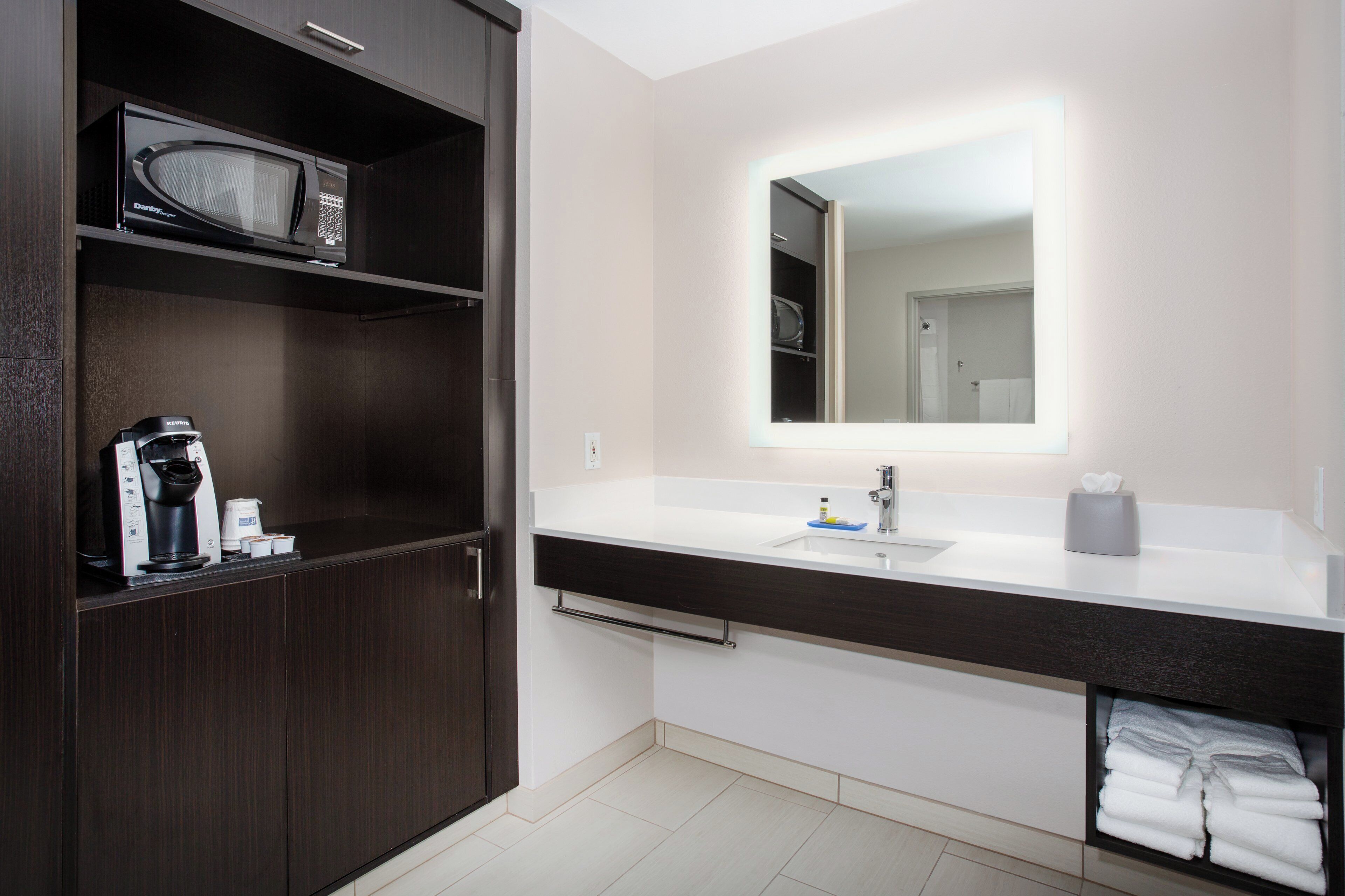 Un baño moderno con tocador de madera oscura, microondas integrado, lavamanos con espejo y un perchero para toallas.