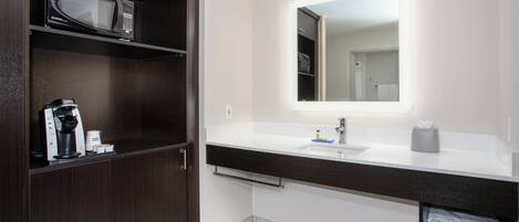 Un baño moderno con tocador de madera oscura, microondas integrado, lavamanos con espejo y un perchero para toallas.
