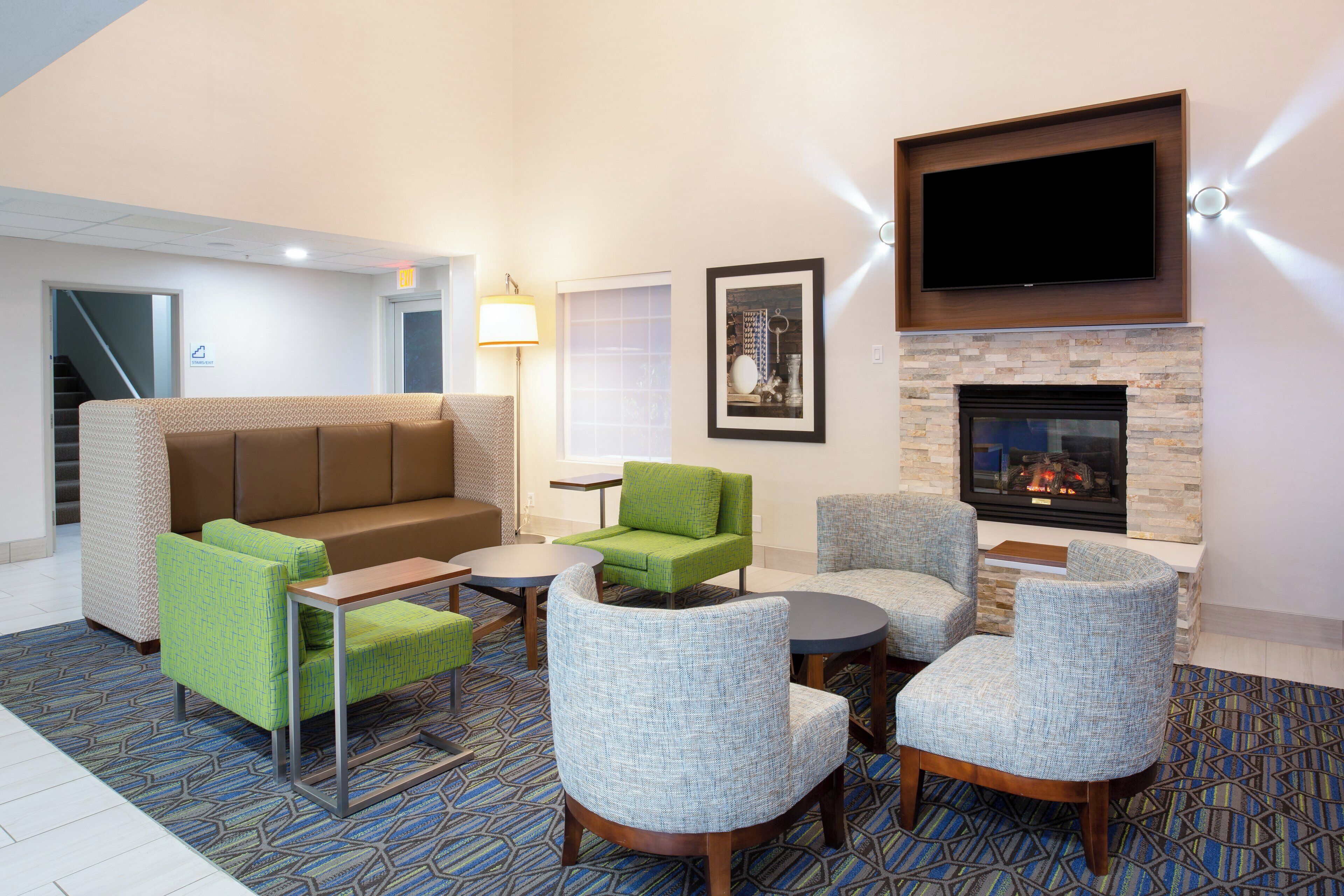 A hotel lobby with a brown sofa, green and blue armchairs, a small table, a fireplace, and a TV mounted above it.