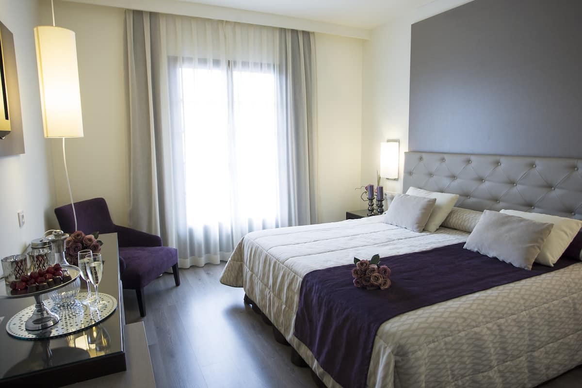 Habitación de hotel con una cama grande, una silla, una mesa con una bandeja de bombones y vasos, y una ventana con cortinas.