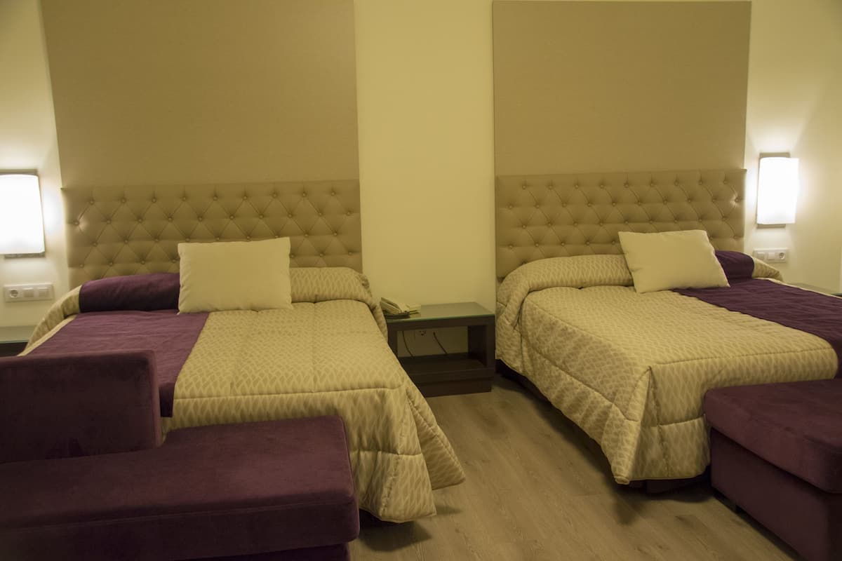 Habitación de hotel con dos camas, cada una con cabecero y mesita de noche. Hay dos sillones morados y una pequeña mesa entre las camas.