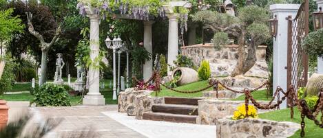 Un sendero en el jardín con escalones de piedra, estatuas y un pérgola con flores colgantes.