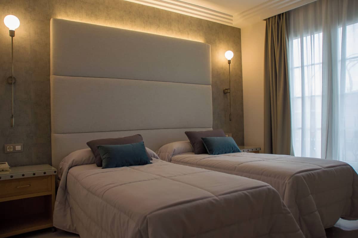 Una habitación de hotel con dos camas, un cabecero grande y acolchado, lámparas de noche y una ventana con cortinas.