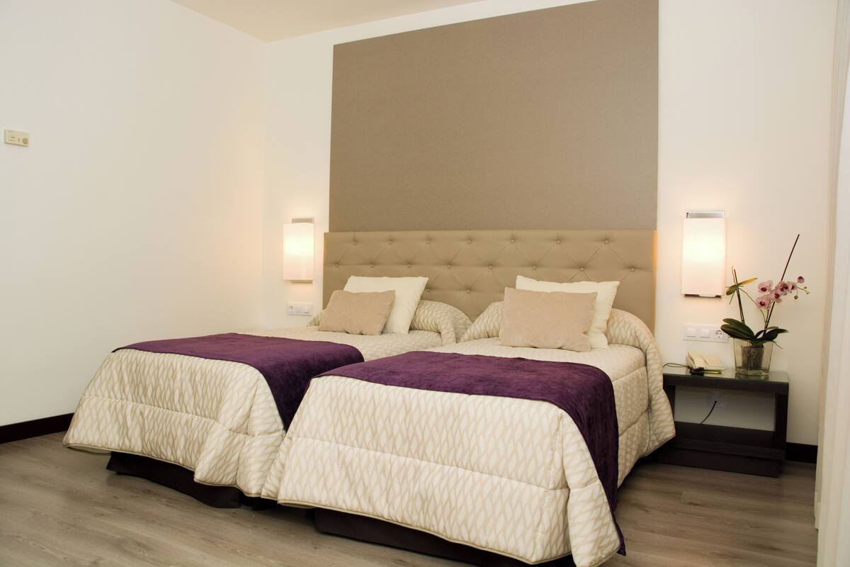 Una cama doble con cabecera capitonada, dos mesitas de noche con lámparas y una mesita auxiliar con un jarrón de flores.