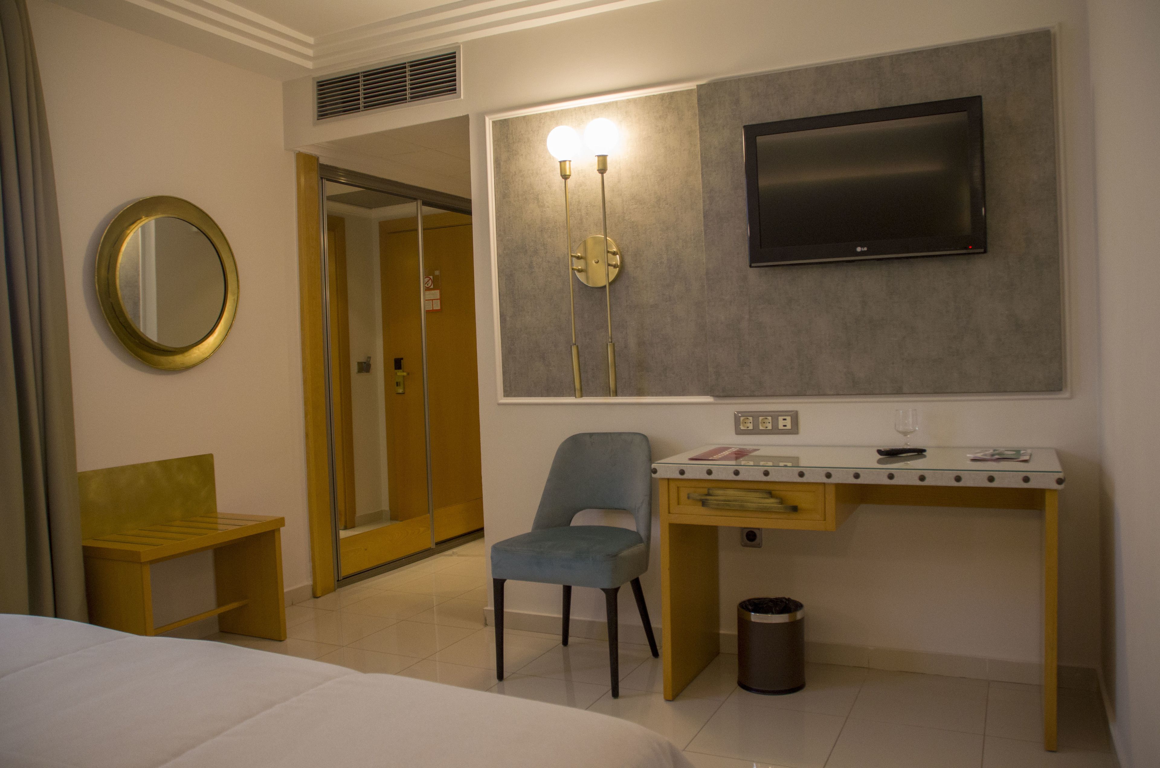 A hotel room with a bed, a desk with a chair, a TV mounted on the wall, a mirror, and a trash can.