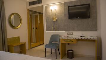 A hotel room with a bed, a desk with a chair, a TV mounted on the wall, a mirror, and a trash can.