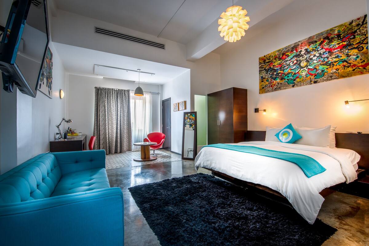 Une chambre d’hôtel moderne avec un grand lit, un canapé bleu, un fauteuil rouge et un tableau abstrait aux couleurs vives accroché au mur.