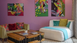 Un salon moderne avec un mur violet, deux tableaux abstraits, un canapé blanc agrémenté de coussins colorés, une table basse en bois avec des livres et des bouteilles, et des carreaux de sol à motifs.