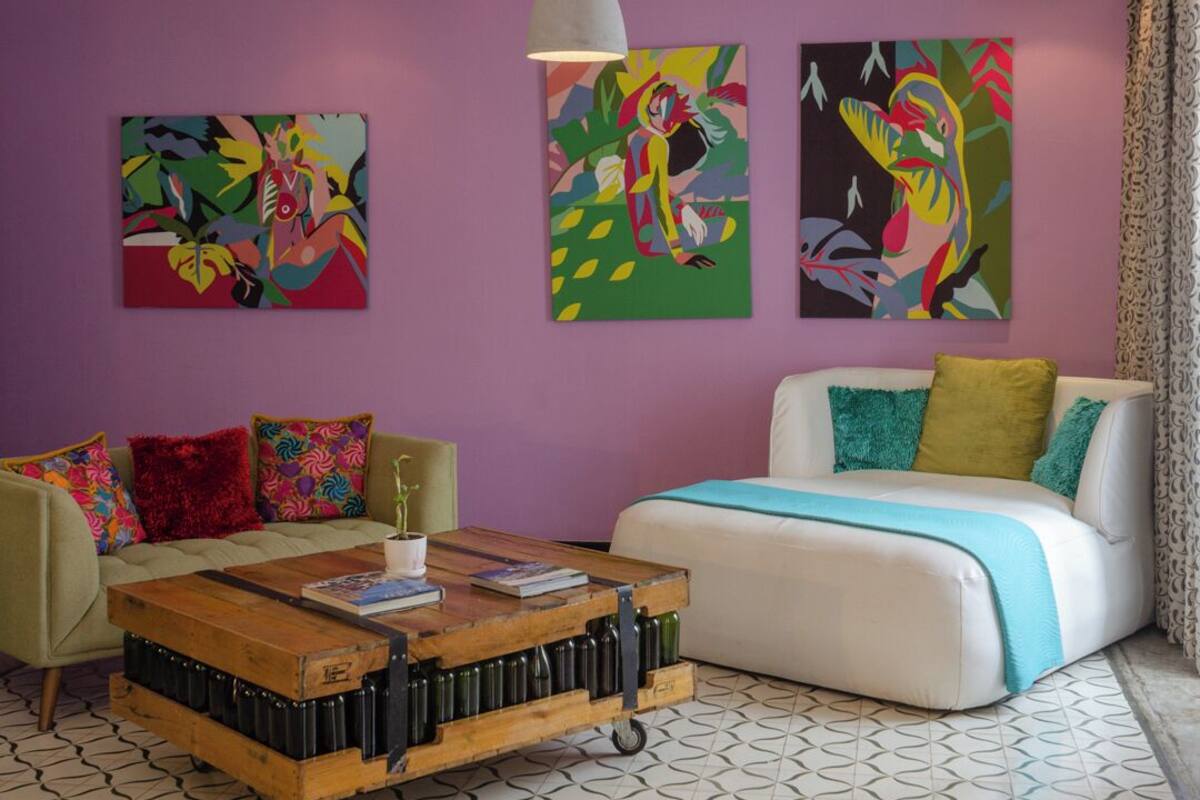 Un salon moderne avec un mur violet, deux tableaux abstraits, un canapé blanc agrémenté de coussins colorés, une table basse en bois avec des livres et des bouteilles, et des carreaux de sol à motifs.