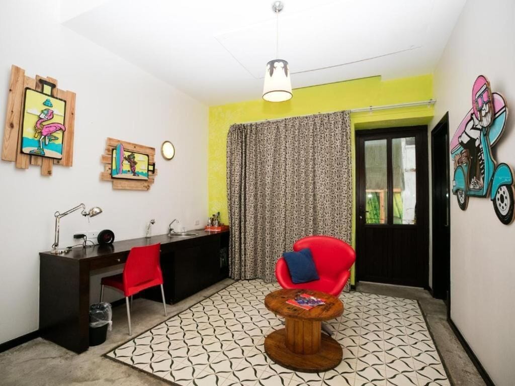 Une pièce comprenant un bureau, une chaise et une petite table ronde. Il y a une fenêtre avec des rideaux, une porte et des œuvres d’art accrochées aux murs.