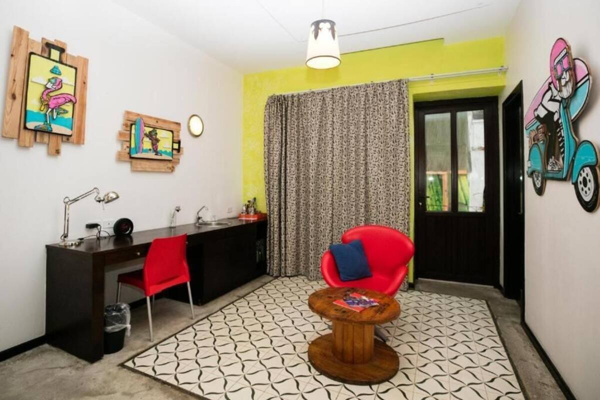 Une pièce comprenant un bureau, une chaise et une petite table ronde. Il y a une fenêtre avec des rideaux, une porte et des œuvres d’art accrochées aux murs.