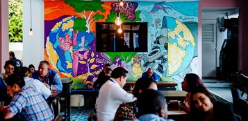 Un mural vibrante con diseños abstractos y una pantalla de televisión en el centro de una habitación con gente sentada en mesas.