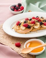 Ein Teller Pfannkuchen, belegt mit Himbeeren, Brombeeren und Bananenscheiben, dazu ein Schälchen Honig und eine kleine Schale mit Beeren.