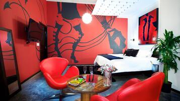 Una habitación de hotel moderna con paredes rojas, una cama, una mesa de comedor con sillas y una planta grande.