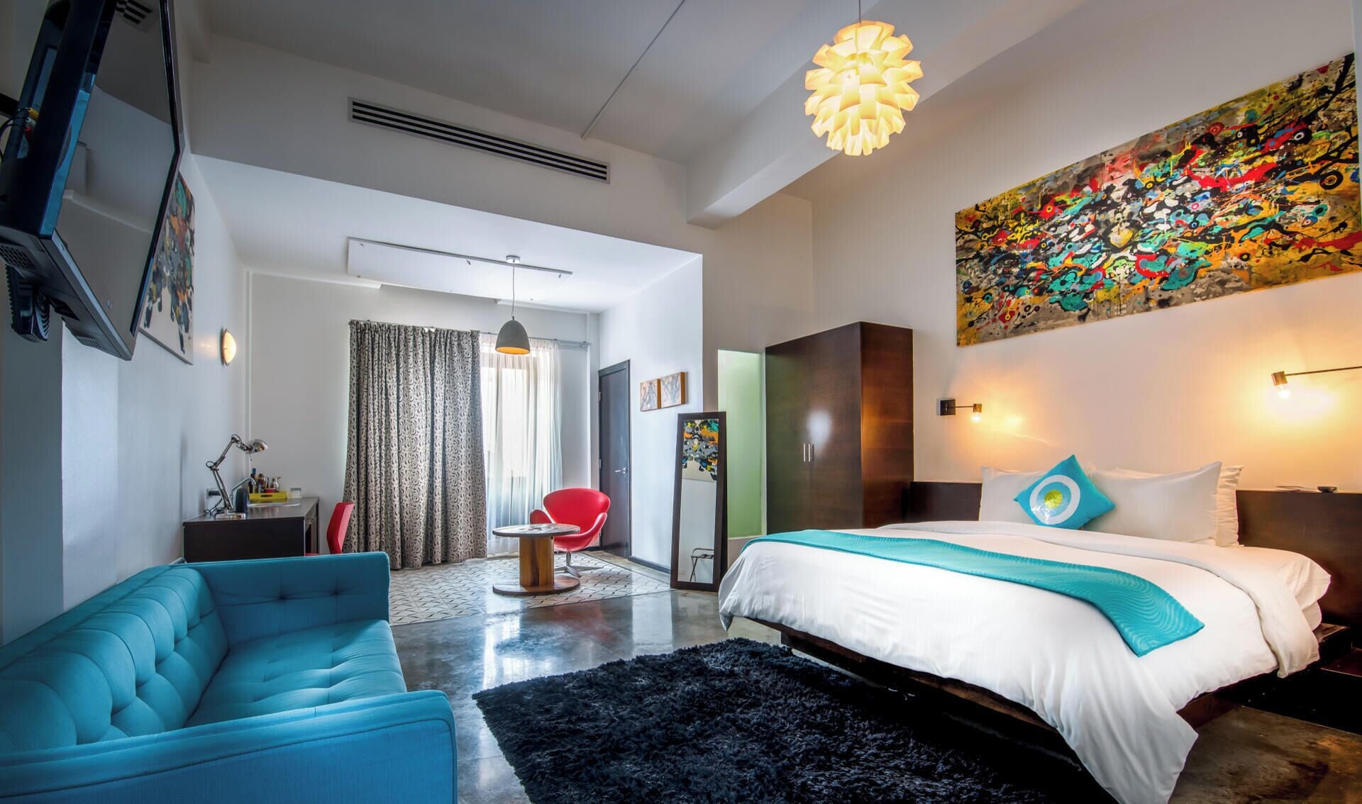 Une chambre d’hôtel moderne avec un grand lit, un fauteuil bleu, une chaise rouge, un tapis noir et une peinture abstraite aux couleurs vives accrochée au mur.