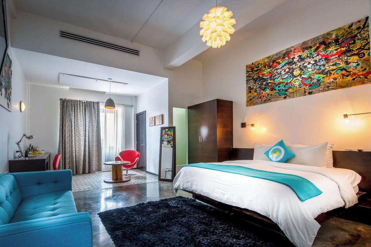 Une chambre d’hôtel moderne avec un grand lit, un fauteuil bleu, une chaise rouge, un tapis noir et une peinture abstraite aux couleurs vives accrochée au mur.