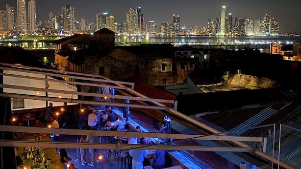Un bar situé sur le toit d’un immeuble, offrant une vue panoramique sur la ville, avec des places assises en plein air et un espace bar où sont exposées des bouteilles et des verres.