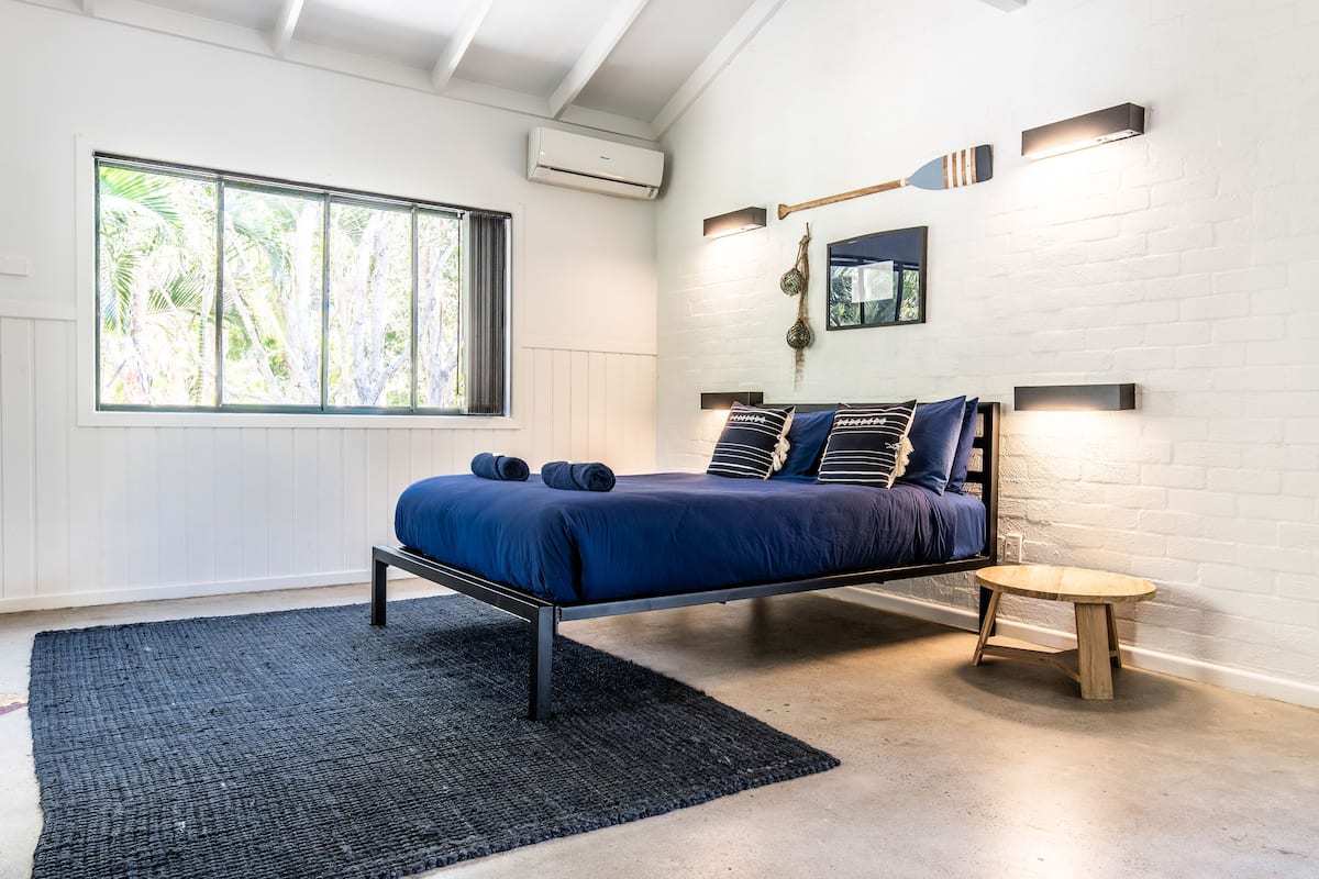 A modern bedroom with a bed, a small wooden table, a large window with a view of trees, and a white brick wall.