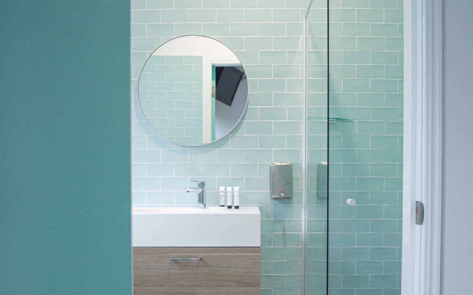 A bathroom with a round mirror, a sink, a glass shower enclosure, and a wooden cabinet.