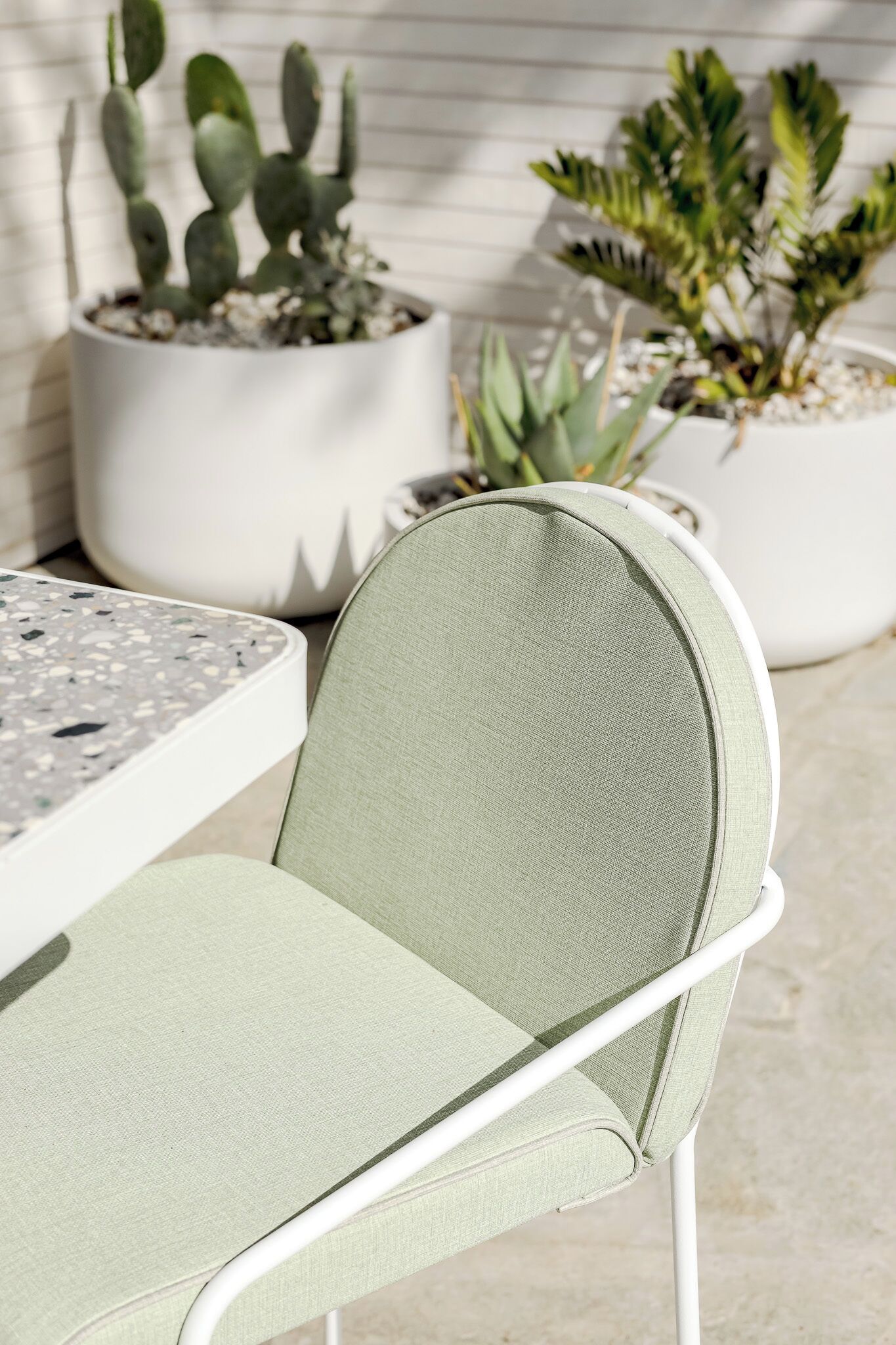 Une chaise avec un cadre en métal et un siège en tissu, une table à la surface mouchetée et, au fond, des plantes en pot.