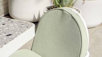 Une chaise avec un cadre en métal et un siège en tissu, une table à la surface mouchetée et, au fond, des plantes en pot.