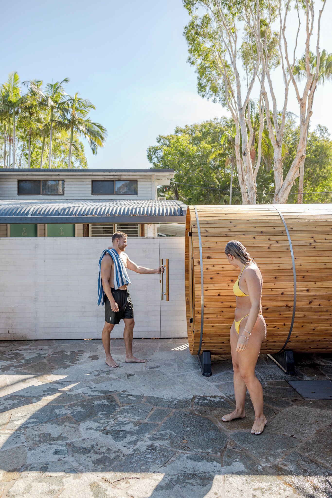 A man and a woman standing near a wooden structure, possibly a sauna or pool, with a building and trees in the background.