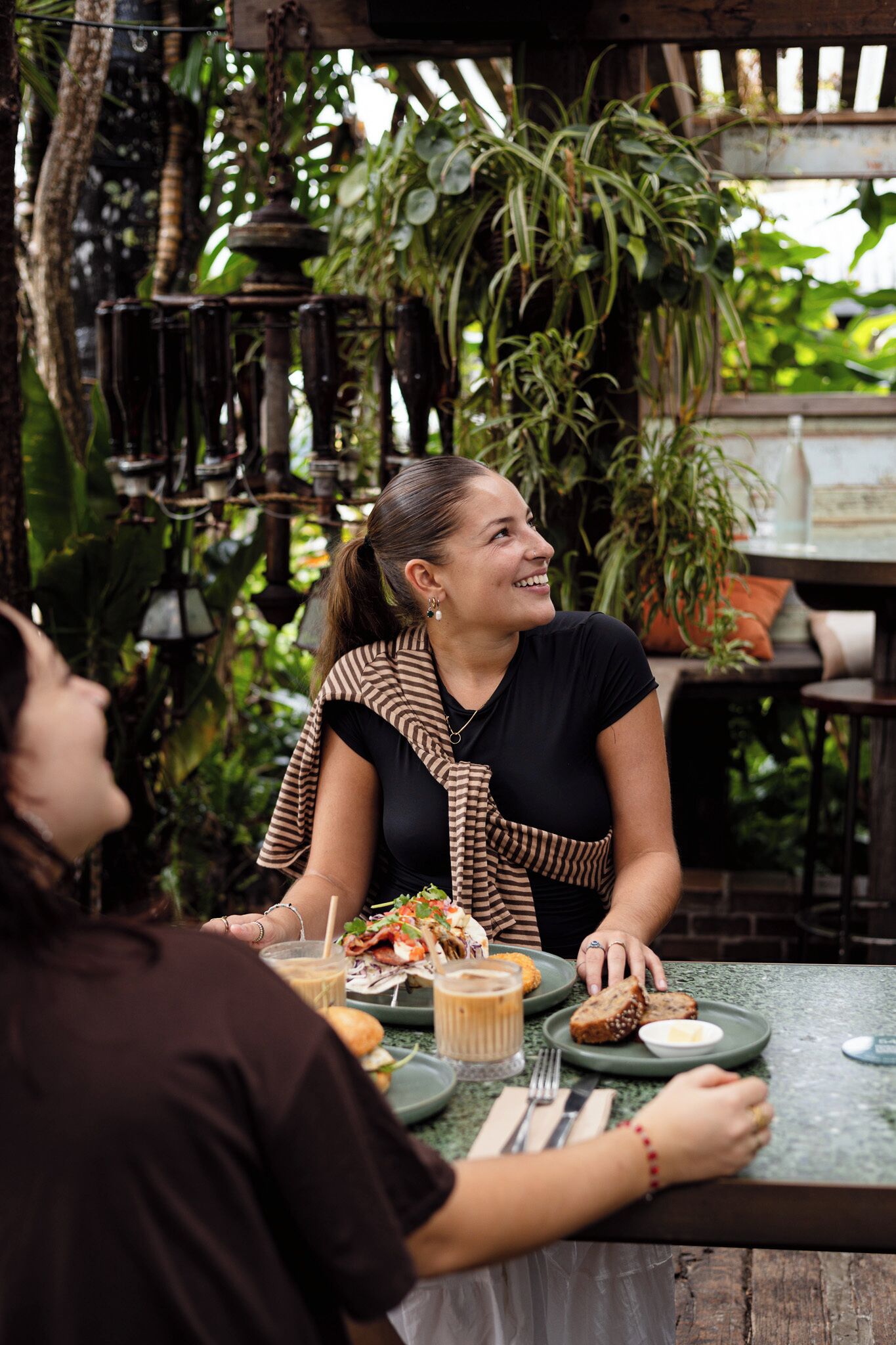 A person seated at an outdoor table with a variety of food items and drinks, surrounded by greenery and plants.
