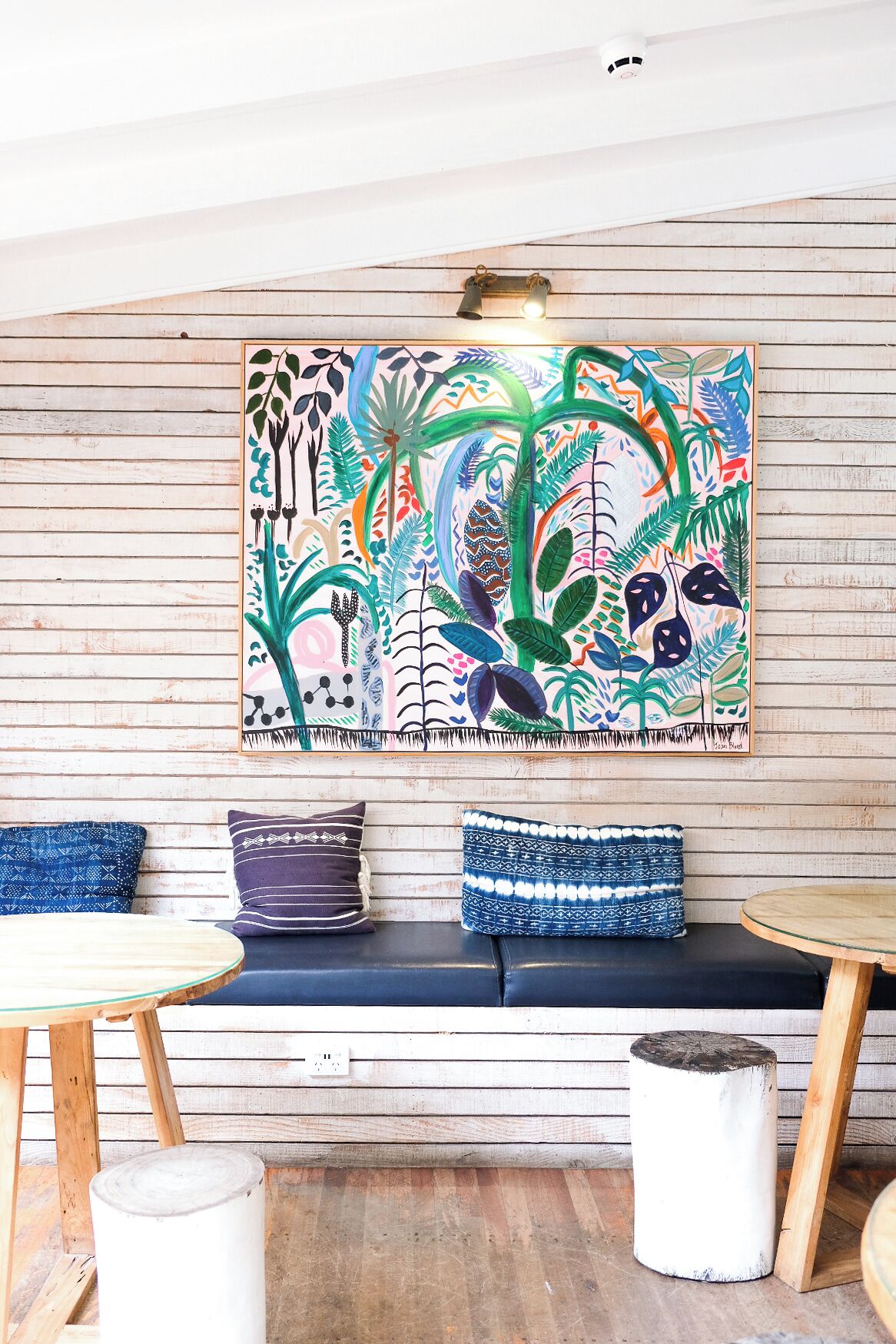 Een wanddecoratie van tropische planten, een houten bank met gestreepte kussens, en een ronde glazen tafel.