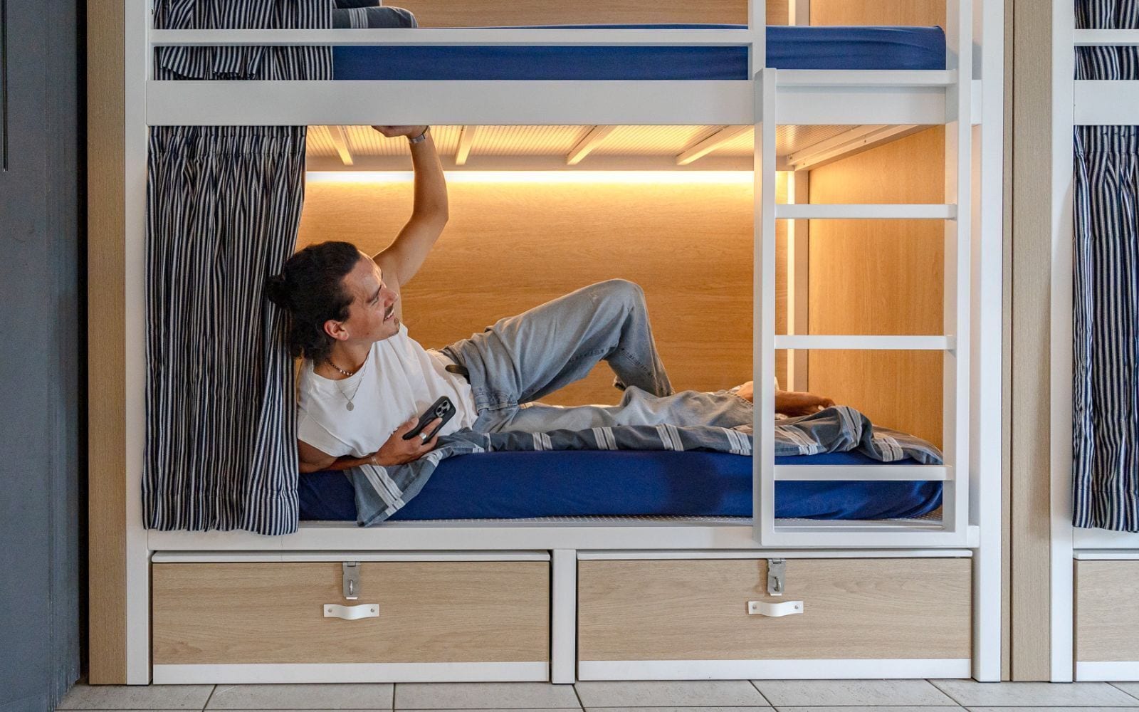 Un lit superposé avec une personne allongée sur le lit du bas, une télécommande et un rideau à rayures.