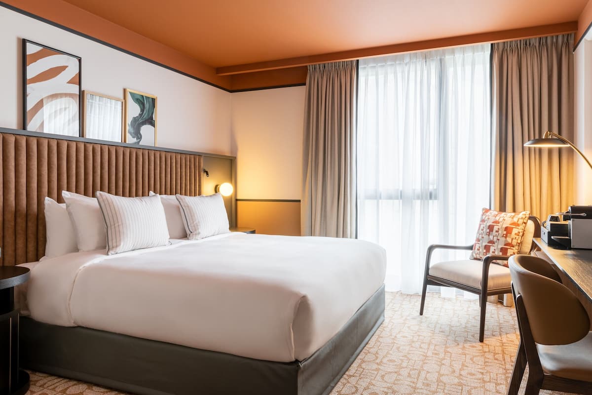 Une chambre d’hôtel avec un grand lit, un bureau et une chaise. Les fenêtres sont dotées de rideaux et un tableau orne le mur.