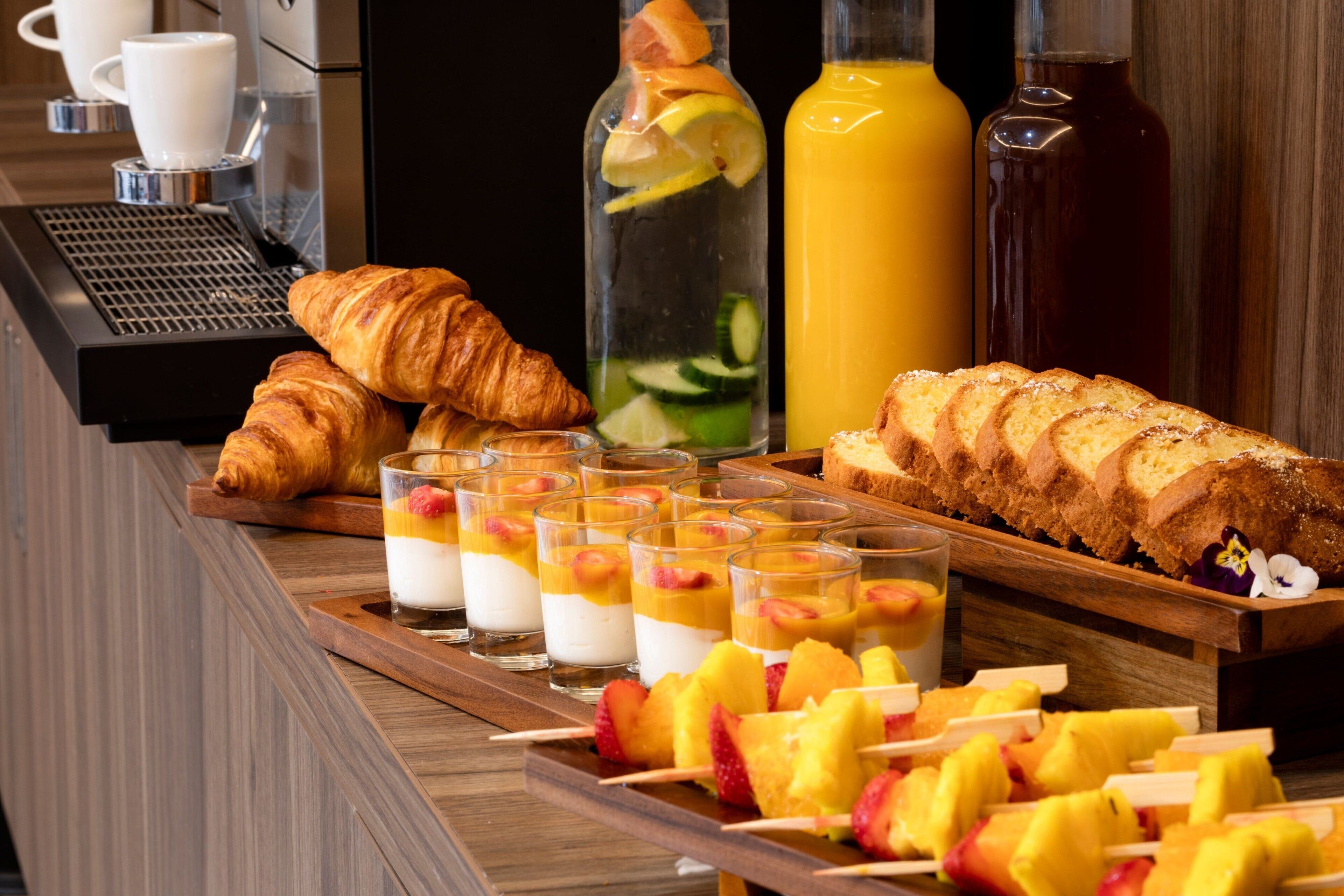 A breakfast buffet with croissants, fruit skewers, parfaits, and juice bottles.