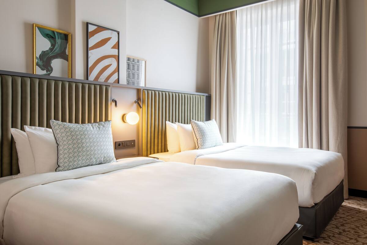 Une chambre d’hôtel avec deux lits, une tête de lit à lattes verticales, une lampe fixée au mur et une grande fenêtre avec des rideaux.