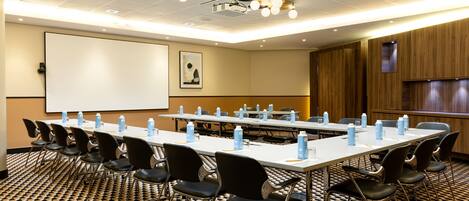 A conference room with a long table, chairs, a projector screen, and a patterned carpet.