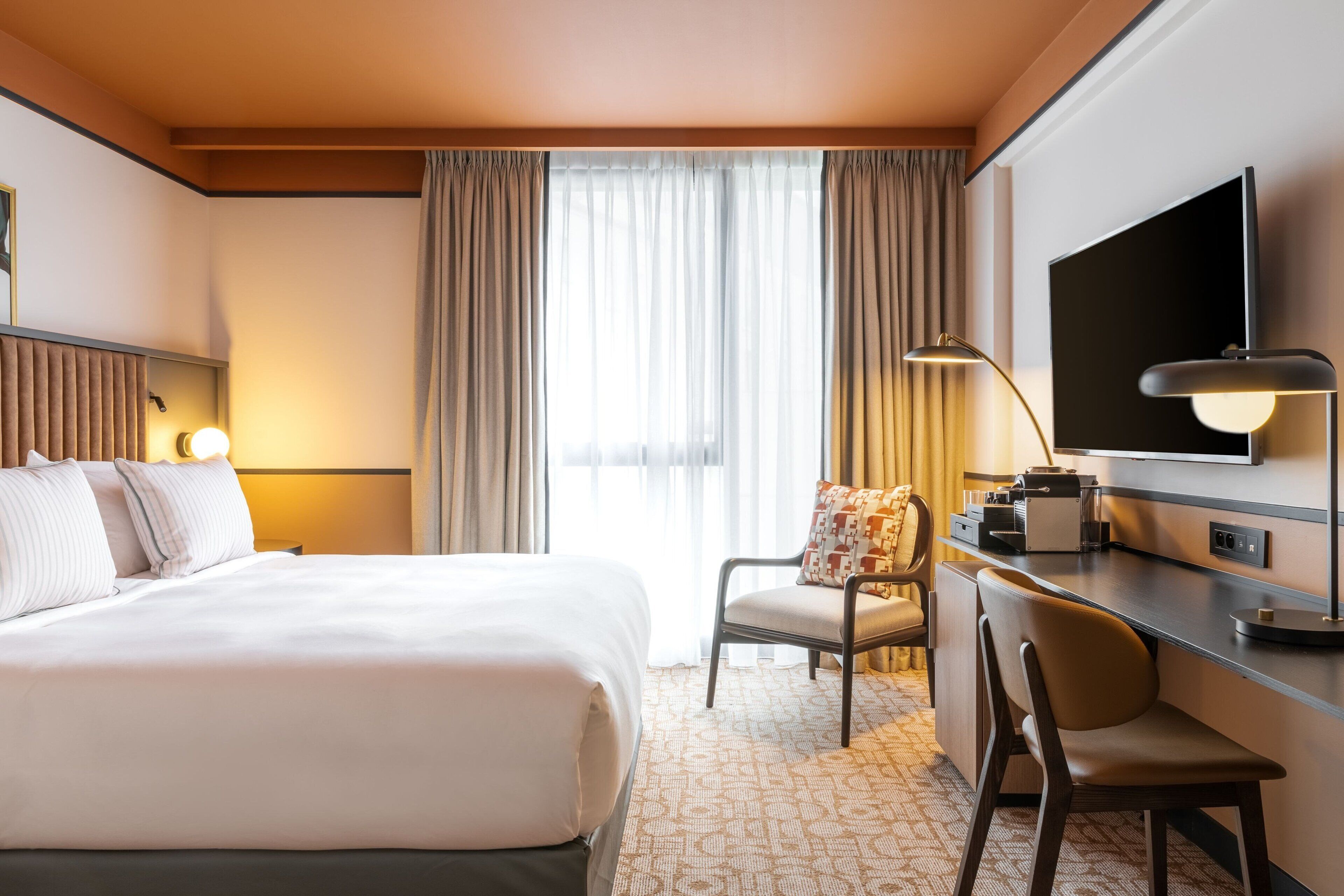 Habitación de hotel con una cama grande, un escritorio con silla, un televisor de pantalla plana y una ventana con cortinas.