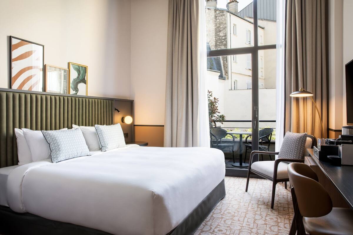 Une chambre d’hôtel moderne dotée d’un grand lit, d’un bureau avec une chaise, d’une vue sur la ville grâce à de grandes fenêtres et d’un balcon avec des sièges d’extérieur.