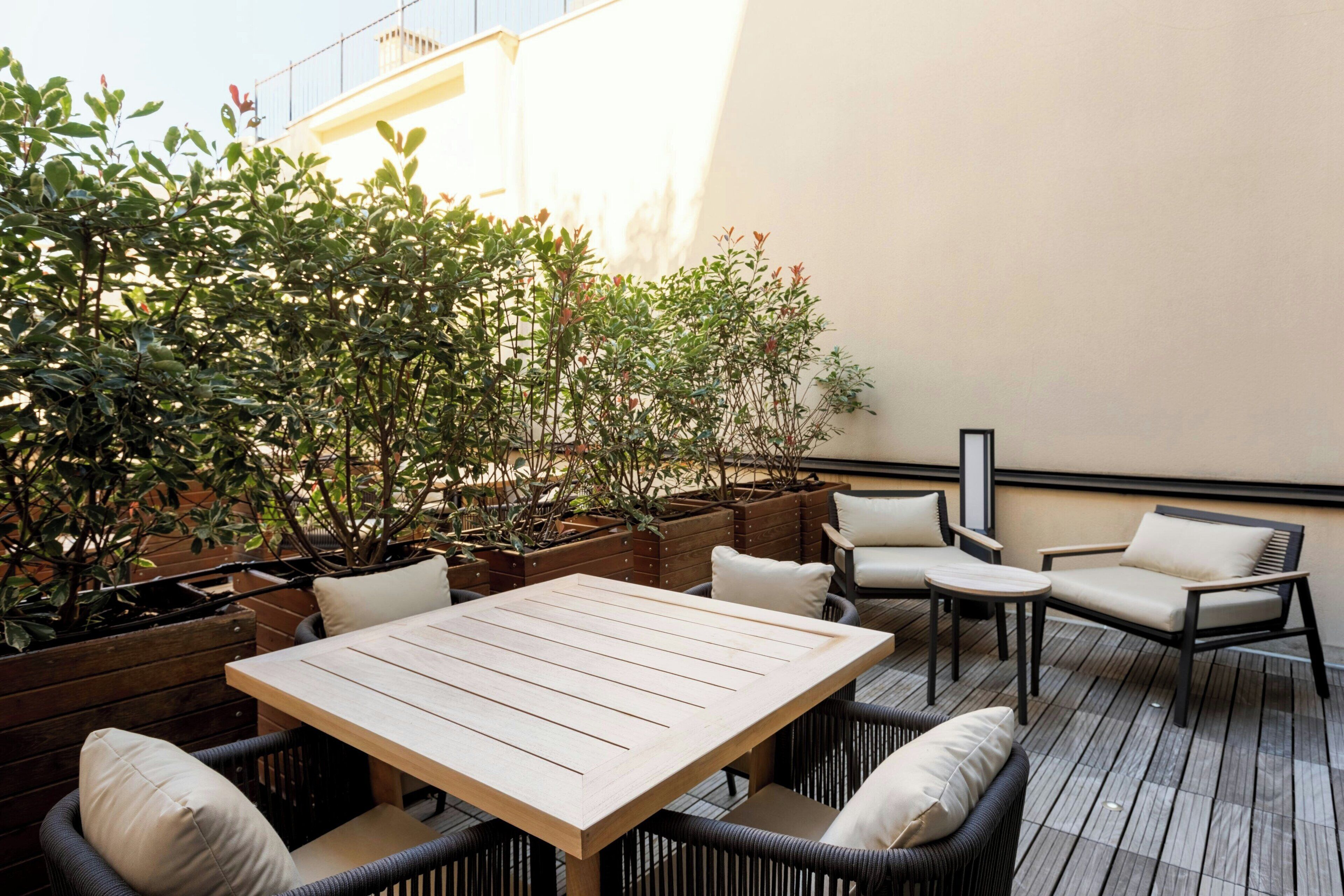 Une terrasse extérieure avec une table en bois et des chaises en osier, entourée de plantes en pot et offrant une vue sur un bâtiment.