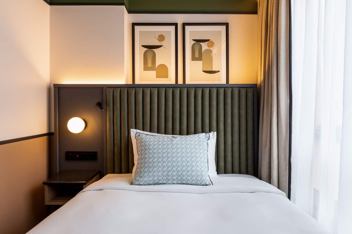 Un lit bien fait, agrémenté d’un coussin à motifs, une lampe fixée au mur et deux œuvres d’art encadrées au-dessus.