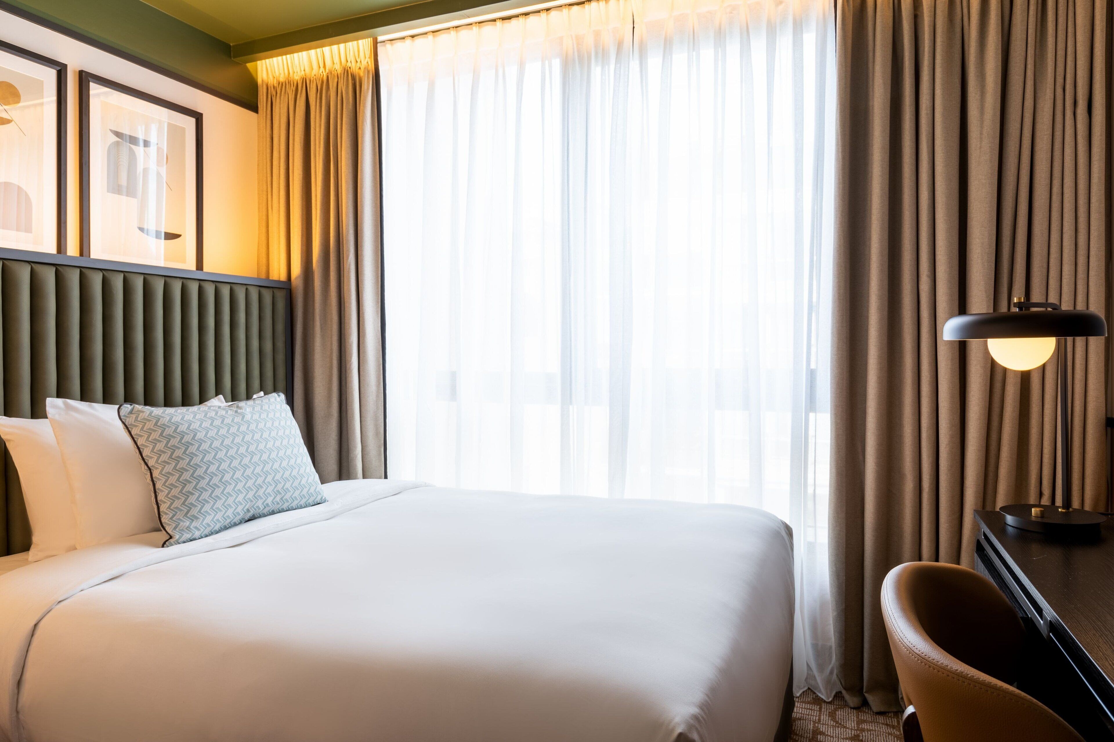 A hotel room with a large bed, a headboard, a desk with a lamp, and a window with curtains.