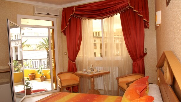 Une chambre d’hôtel avec un lit, deux chaises, une petite table avec un verre, et une vue sur le balcon extérieur.
