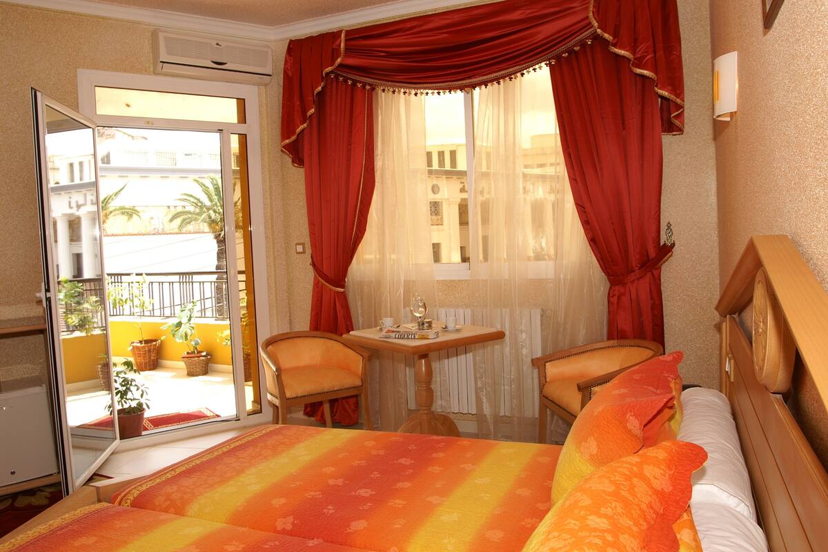 A hotel room with a bed, two chairs, a small table with a glass, and a view of the outside balcony.