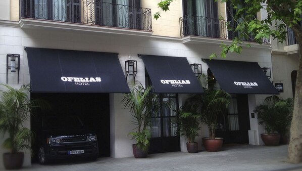 A fachada do Hotel Oferias com três toldos pretos, um Range Rover preto estacionado em frente e plantas em vasos decorando a entrada.