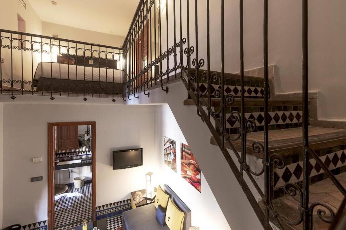 Un intérieur à plusieurs niveaux avec un escalier, un espace de vie comprenant un canapé et un téléviseur, et une cuisine visible à travers une porte ouverte.