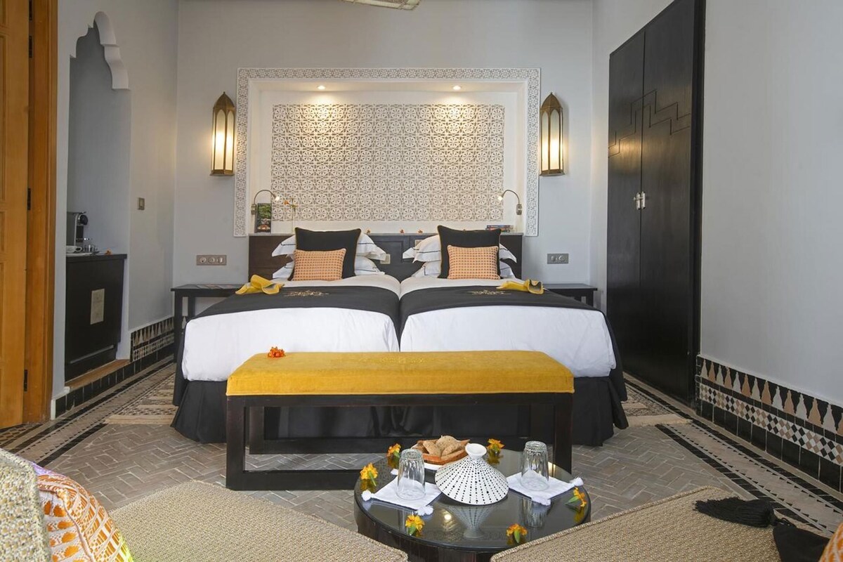 A hotel room with two beds, a small table with a yellow bench, and a decorative wall panel.