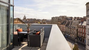 A rooftop terrace with a table and chairs, a potted plant, and a view of historic buildings.